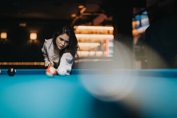 A young female player lines up her shot at a pool table in a dark recreation room, showcasing...