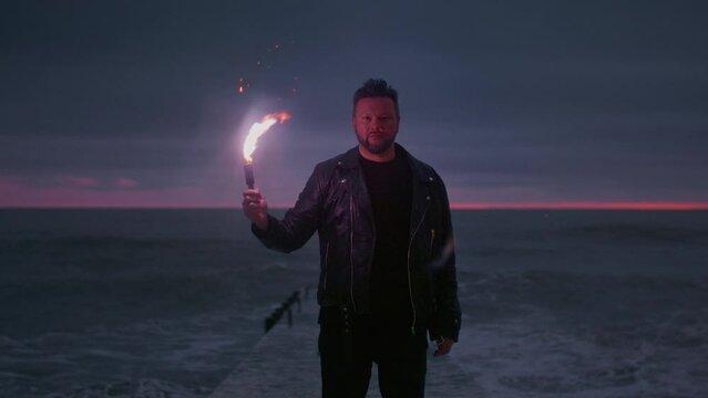 Portrait: a man traveler in a leather jacket holds a red signal light in his hands. Male stands on the breakwater, burning a fire stick or torch. Sea or ocean in the evening, big wave. Slow motion