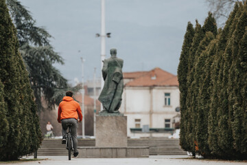 A young man in a red jacket rides his bicycle through a serene park, with overcast weather adding a...