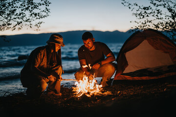 Two friends gather around a glowing campfire near a lake, preparing food as dusk settles,...