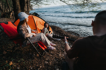 Group of friends gathered by a lake, relaxing and having fun at a campsite with a tent and chairs