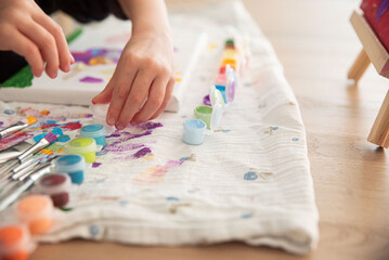Cute little child painting with paintbrush and colorful paints. Kid hands start painting at the table with art supplies, top view