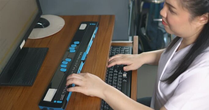 Asian woman with blindness disability using computer with refreshable braille display or braille terminal a technology assistive device for persons with visual impairment in workplace, handheld shot.
