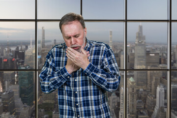 Sad mature caucasian man is coughing standing indoors. Windows background with cityscape view.