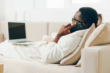 African American man working on laptop in his cozy home, wearing a bathrobe and glasses, with a phone next to him He's enjoying his morning, chatting and making online business calls while sitting on