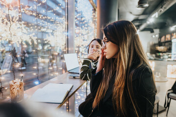 Two young professionals collaborate on a project in a warmly lit cafe adorned with sparkling festive lights, exuding a cozy, creative, and focused atmosphere.
