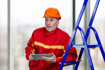 Portrait of elder caucasian contruction worker is using tablet device. Man in special work uniform lean on a blue ladder.
