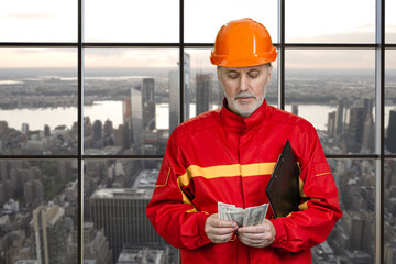 Portrait of construction worker in red uniform is counting money. Urban cityscape background.