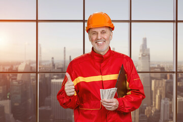 Portrait of happy elder man holding money and showing like gesture. WIndows with view of urban cityscape background.