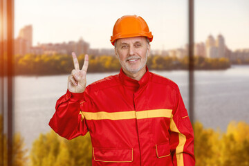 Happy smiling construction worker shows victory gesture sign. Window with river scape background.