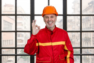 Smiling construction handyman worker in red uniform shows okay gesture sign. Checkered window background.