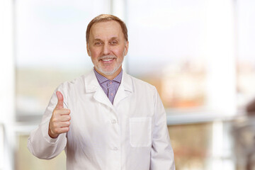 Happy male senior medic in white coat shows thumb up. Abstract indoor window background.