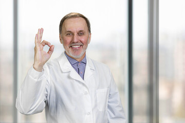 Cheerful senior doctor shows okay gesture sign. Everything is fine. Blurred window in the backgroud.