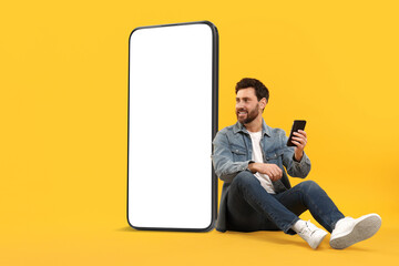 Man with mobile phone sitting near huge device with empty screen on orange background. Mockup for...