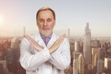 Happy senior medic doctor in white coat shows no reject sign. Urban cityscape background.