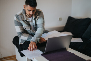 A young male professional concentrates on his work, using a laptop at home while analyzing documents.