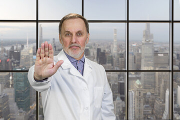 Portrait of senior medic doctor in white coat shows no reject sign by his hand. Checkered window background with urban scape view.