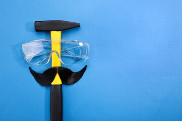Man's face made of artificial mustache, safety glasses and hammer on blue background, top view....
