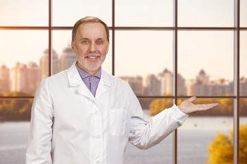 Happy smiling male senior doctor showing a copy space. Checkered window background with landscape view.