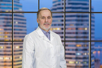 Portrait of senior doctor with night cityscape background. Smiling confident physician.