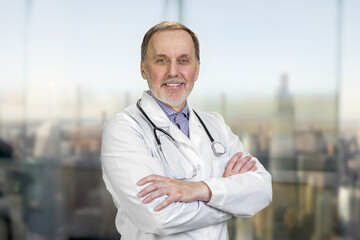 Portrait of smiling senior doctor with folded arms. Blurred cityscape background.