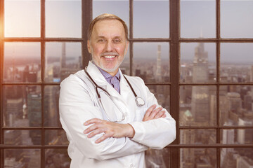 Portrait of happy senior doctor with folded arms. Evening cityscape view.