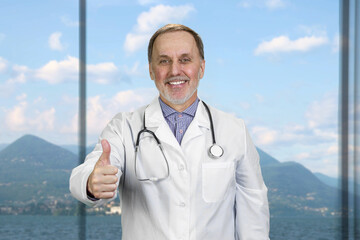 Happy smiling senior doctor with stethoscope shows thumb up. Window with landscape view in the background.