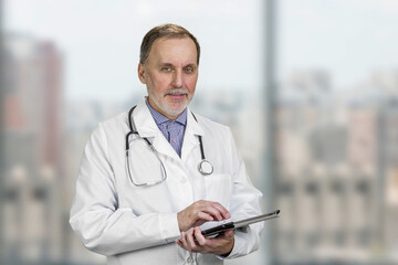Portrait of mature senior male doctor holding digital tablet standing indoors. Abstract blurred windows background.