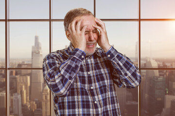 Portrait of a handsome senior mature male holds his head with both hands. Checkered windows background with cityscape view.