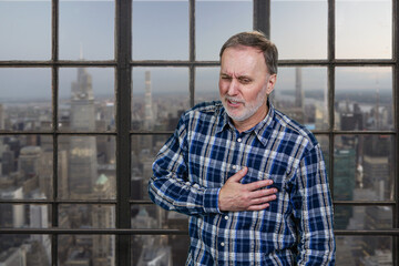 Portrait of a mature man having heartache touching his chest. Aged man having heart atack indoors. Checkered windows background with cityscape view.