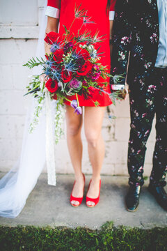 Photo of Bride Wearing Mini Red Wedding Dress and Long Veil Holding Hands with Groom in Floral Wedding Suit, Textured Red Rose Wedding Bouquet