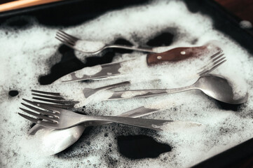 A knife with wooden handle, fork and spoons in the detergent foam on a black oven-tray. Washing dishes concept.
