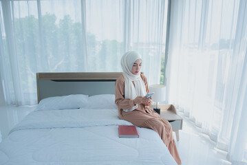 Asian Muslim woman sitting on white bed and reading the Qur'an of Islam that Muslims believe is...