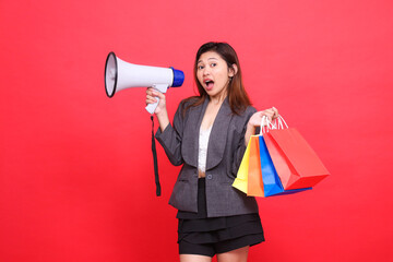 Asian sales woman shouts cheerfully at the camera through a megaphone speaker and holds up a shopping paper bag wearing a gray jacket on a red background. for transaction, lifestyle and business