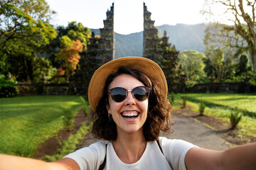 Happy young woman traveler looking at camera taking selfie in Balinese Hindu Temple entrance during...