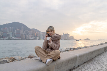 Fototapeta na wymiar A young woman in winter clothes in her 20s who eats local food fish balls in the evening in a park with a view of buildings and the sea on Kowloon Island, Hong Kong 香港九龍島のビル群と海が見える公園で夕方にローカルフードのフィッシュボ