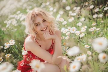 A beautiful young woman in a red dress posing for the camera, smiling and holding daisies in her hands while sitting amidst tall grasses covered with white wildflowers.