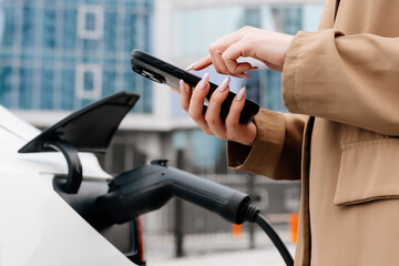 A business woman uses an electric charging station for her modern car against the backdrop of a modern city. Concept of economic charging and modern technologies.