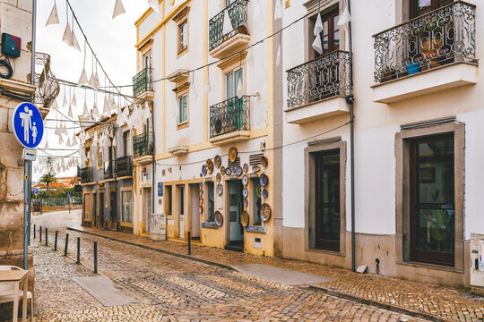 Street in Tavira, Portugal, with cobblestone pavement, hanging decorations, and traditional white buildings with iron balconies.