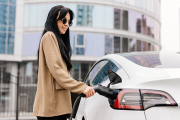 Obraz premium Close-up of a modern electric car charging against the backdrop of a business woman and a modern business center. Concept of using energy, electrical machines and modern technologies.