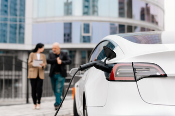 An unrecognizable modern electric car is charging. In the background, two business people, a man...