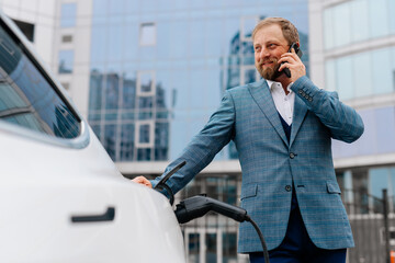 A middle-aged businessman charges an electric car from a charging station powered by renewable...