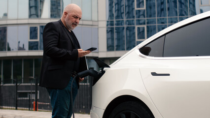 A business man uses a mobile phone while charging the battery of his electric car. A confident man in a business suit stands near a luxury car with a modern smartphone in his hands. 