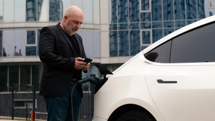 A middle-aged businessman charges an electric car from a charging station powered by renewable...