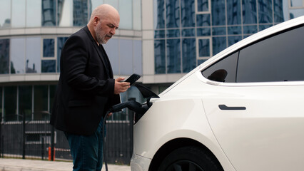 A businessman turns off an electric car with a full charge level at a charging station using an...