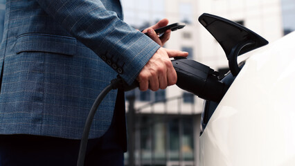 A businessman turns off an electric car with a full charge level at a charging station using an...