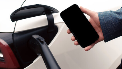Close-up photo of an electric car charging. A man is holding a phone with a black screen for your...