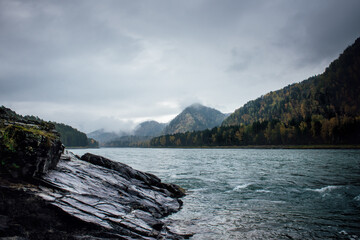 The stone shore of a large and fast mountain river surrounded by mountains covered with mixed autumn forest. A cold autumn morning in the wild with thick low clouds