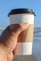 personal view of hand holding disposable cup of coffee on beach