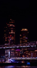 Obraz premium Millennium Bridge illuminated at night, with tourists walking and a night train stopped on Blackfriars Bridge with skyscrapers behind with illuminated windows and the London Eye in the background.